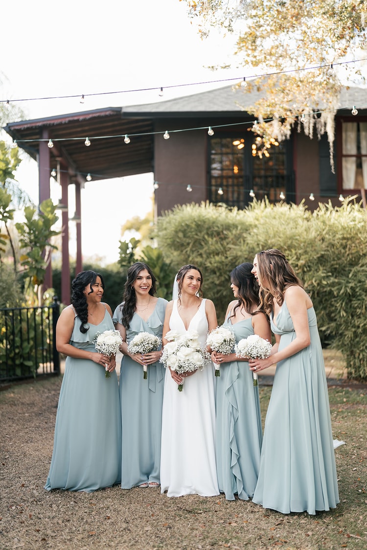 Bride surrounded by her four bridesmaids in sage dresses, all holding white bouquets and laughing together in the garden.