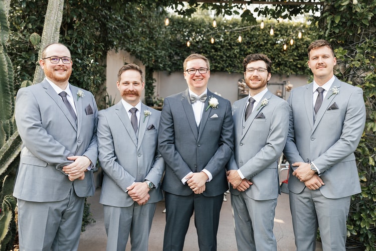Groom posing at center with four groomsmen in light gray suits, all wearing boutonnieres and smiling under a leafy overhead structure.