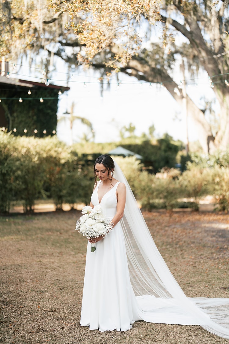Bride standing alone in the garden holding her bouquet, cathedral veil trailing behind her, looking down with quiet focus.
