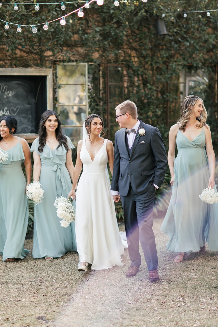 Wedding party walking together through the venue grounds, bride and groom hand in hand at center, bridesmaids in sage dresses alongside.