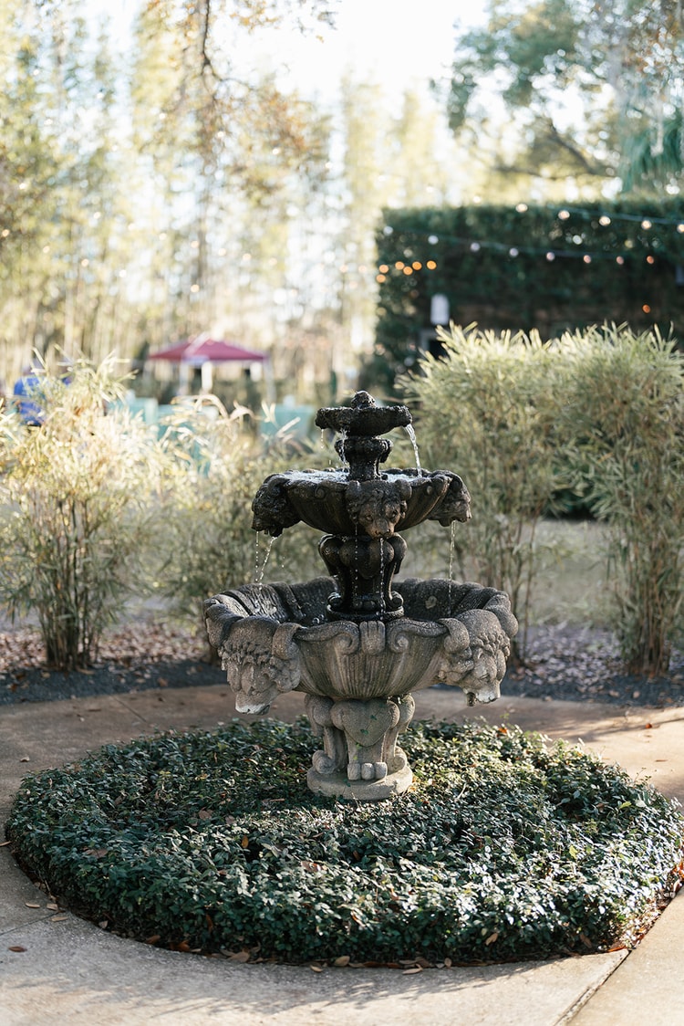 Stone tiered garden fountain flowing amid lush greenery and string lights at the venue.