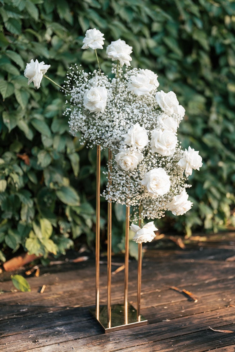 Tall floral arrangement of white roses and baby's breath on a gold pillar stand, placed against an ivy-covered garden wall.