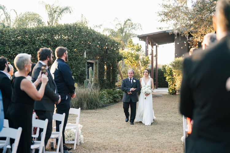 Bride being walked down the aisle by a family member at sunset, guests standing and watching as they approach the altar.