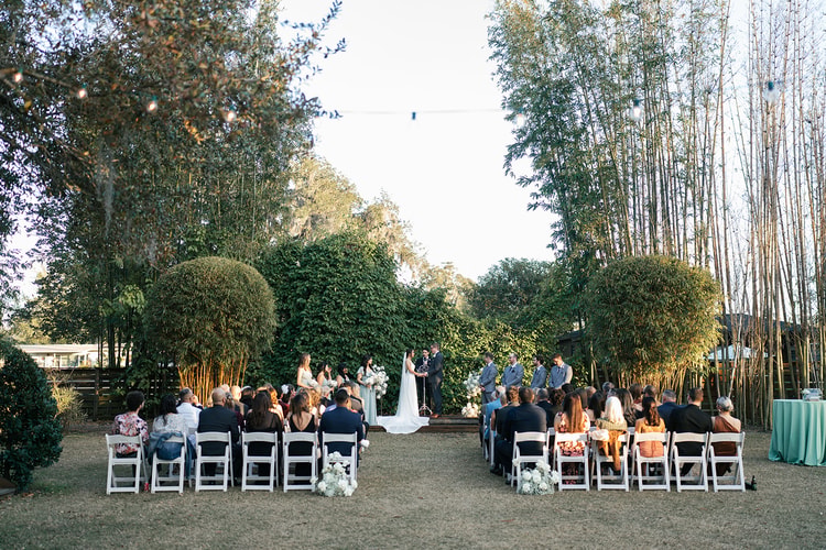 Wide view of the outdoor ceremony from behind the guests, couple at the altar surrounded by tall greenery and soft evening light.