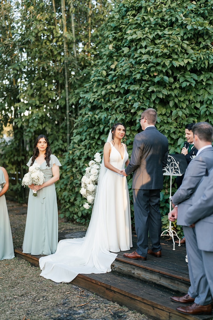 Bride and groom facing each other at the altar during the ceremony, officiants beside them, bridesmaids standing to the left.