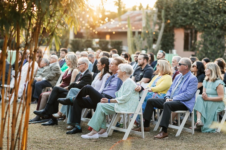 Guests seated in white folding chairs watching the outdoor ceremony at sunset, string lights woven through the trees above.