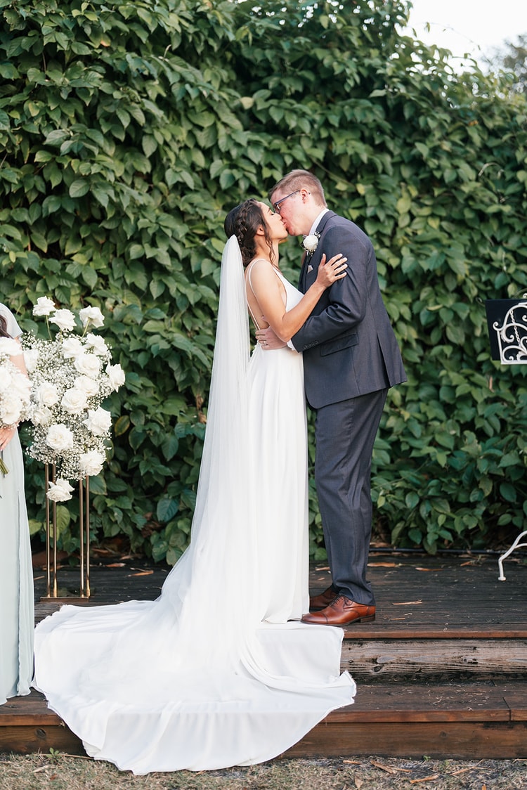 Bride and groom sharing their first kiss at the altar, veil flowing behind her, white floral arrangements framing the moment.