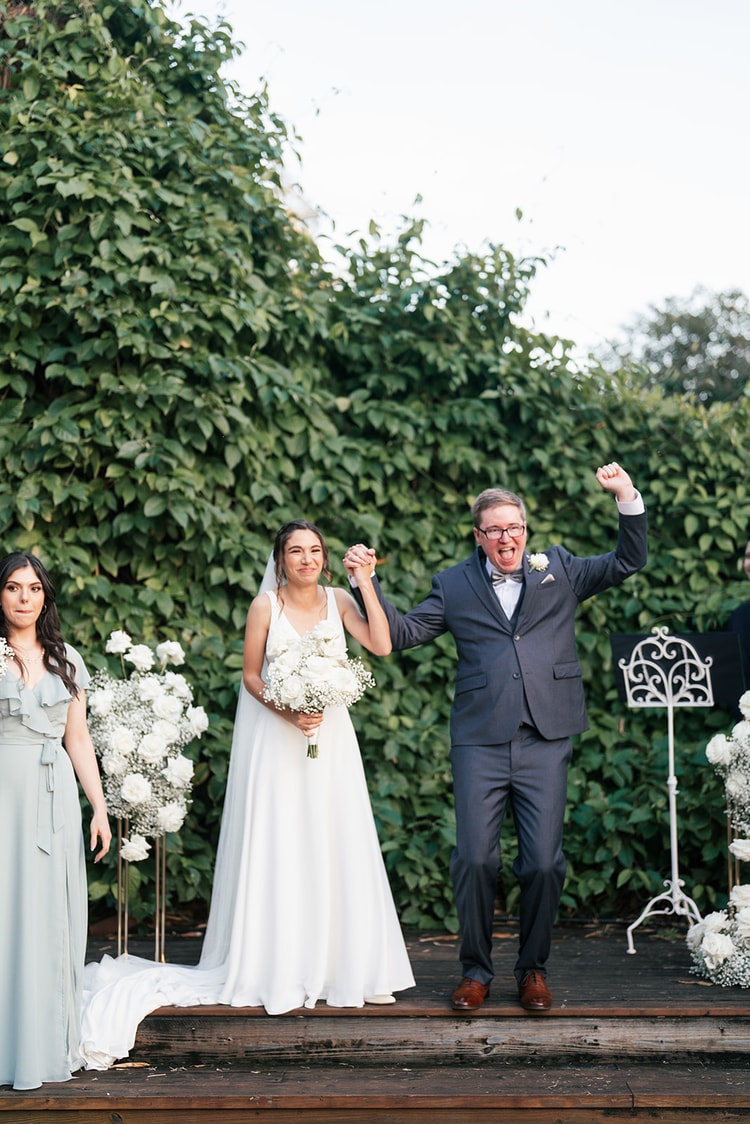 Couple at the altar, groom pumping his fist in celebration as the officiant announces them married, bride laughing joyfully.
