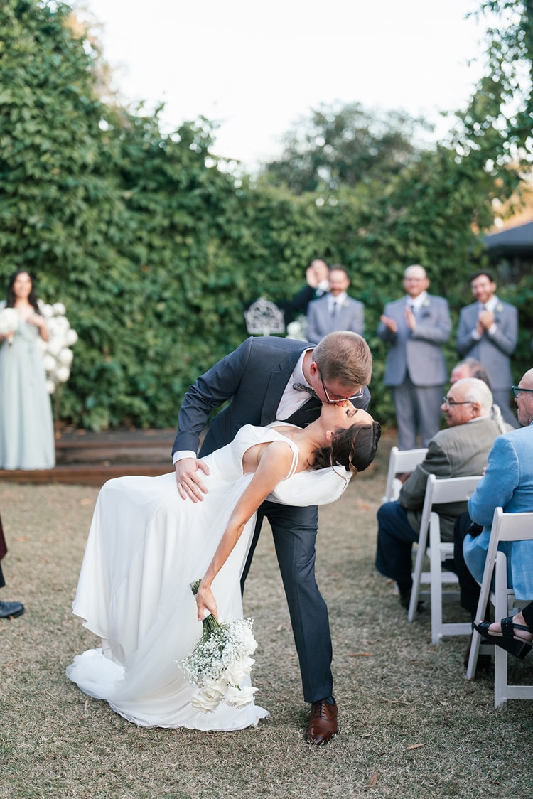 Groom dipping the bride for a celebratory kiss after the ceremony recessional, wedding party and guests applauding behind them.