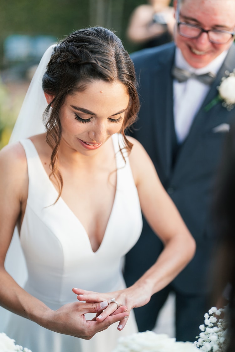 Bride gazing down at her new wedding ring just after the exchange of vows, groom beaming in the background.