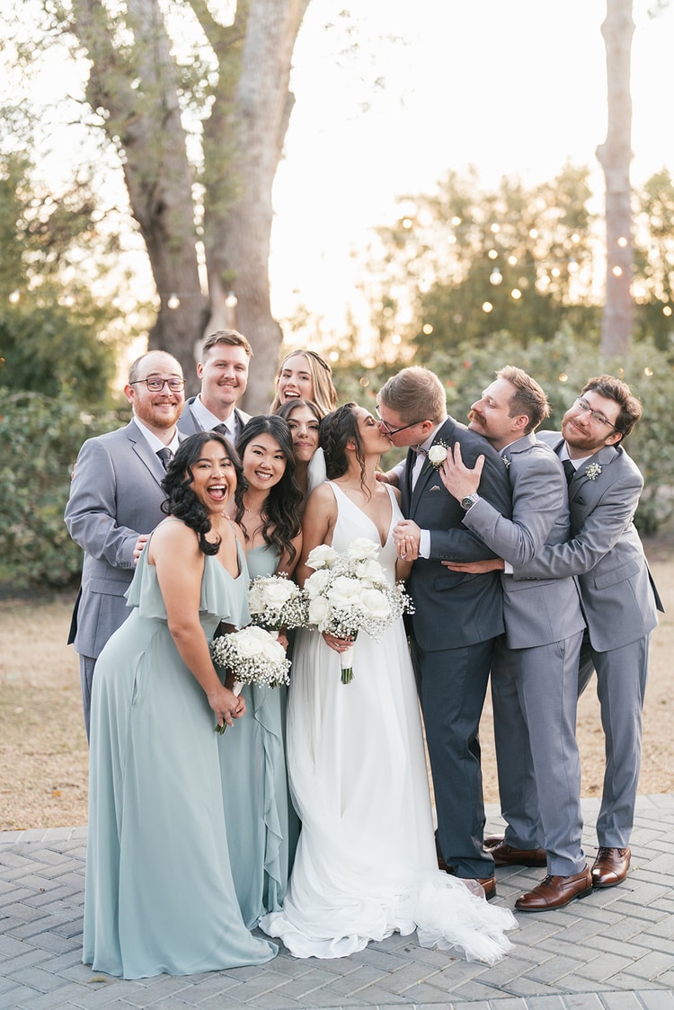 Full wedding party grouped together for a fun portrait at golden hour, groom kissing the bride at center while everyone laughs and cheers.