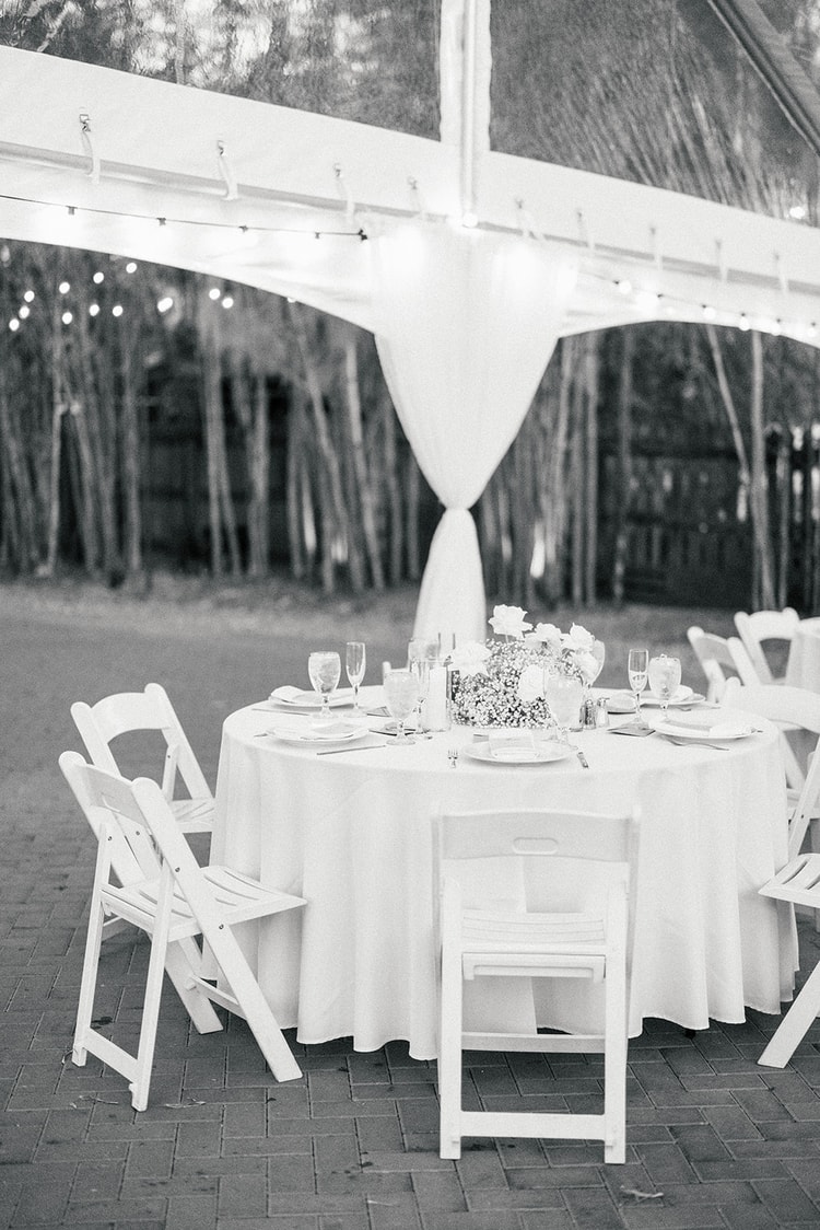 Reception table under the clear tent at night, white linen and floral centerpiece, draped fabric column in the background, black and white.