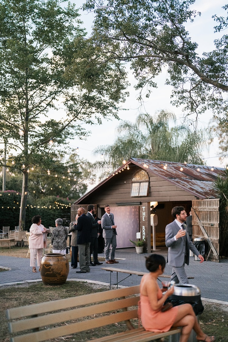 Cocktail hour scene at dusk with guests mingling near a barn structure and trees strung with Edison bulb lights.