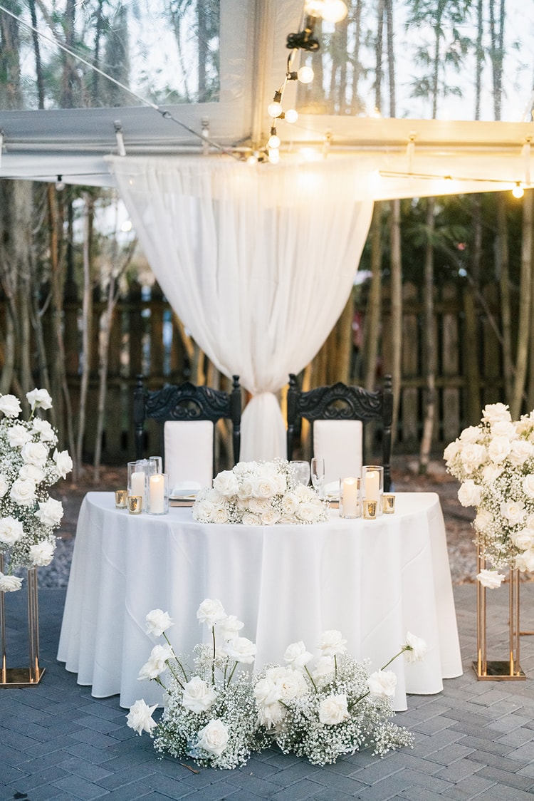 Sweetheart table set with white roses, baby's breath, and candles beneath draped fabric and string lights, flanked by tall gold floral pedestals.