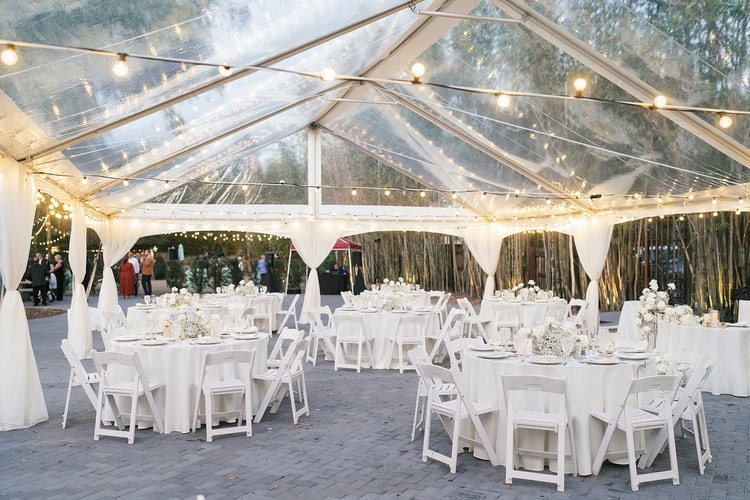 Reception tent interior with a clear roof, string lights overhead, and white-draped round tables set for dinner.
