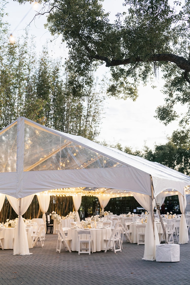 Reception tent exterior at dusk, string lights glowing through the clear canopy, white tables visible through the open sides.