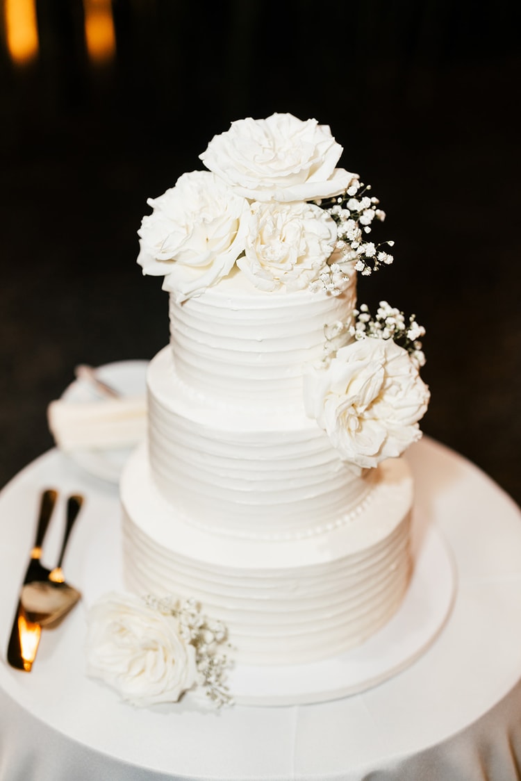 Three-tier white buttercream wedding cake decorated with white roses and baby's breath, gold cake server beside it.