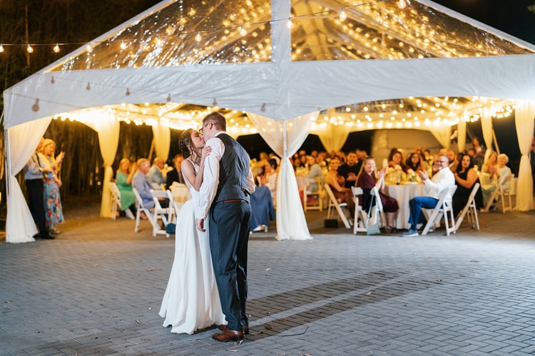 Bride and groom sharing a kiss on the open-air dance floor at night, guests seated at illuminated tables under the tent behind them.