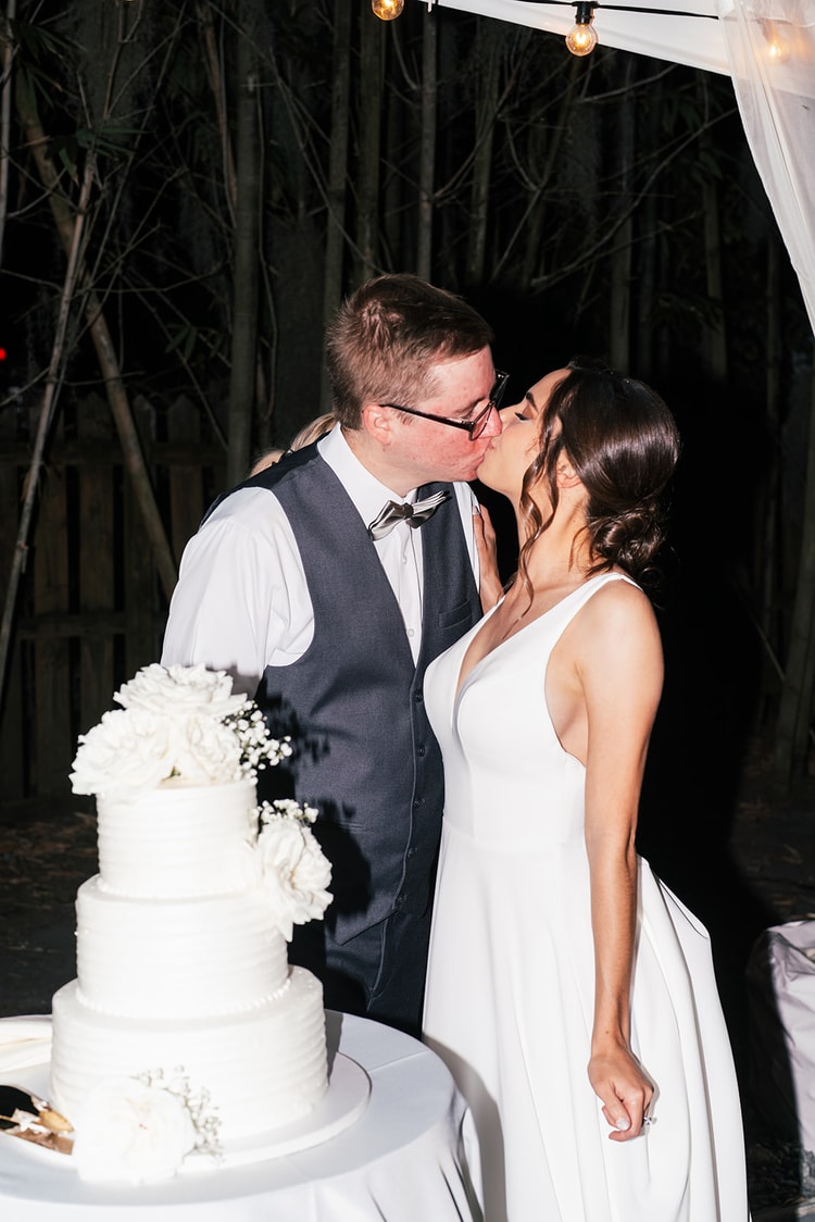 Couple kissing beside the wedding cake at night reception, groom in vest and bow tie, cake topped with white florals in the foreground.