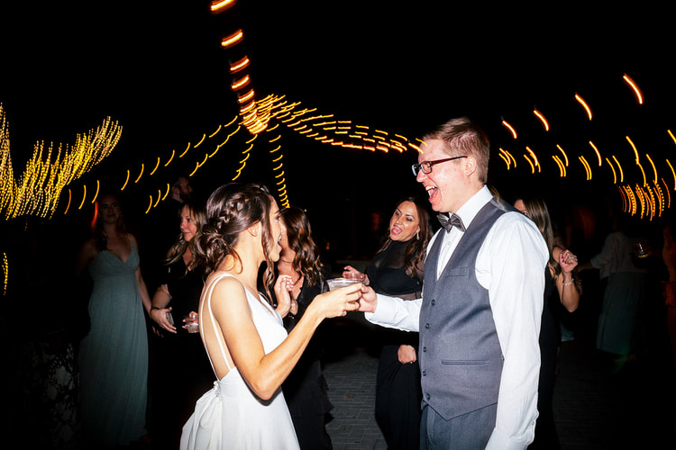 Bride and groom dancing and laughing together on the crowded reception dance floor, warm light trails spinning through the dark behind them.