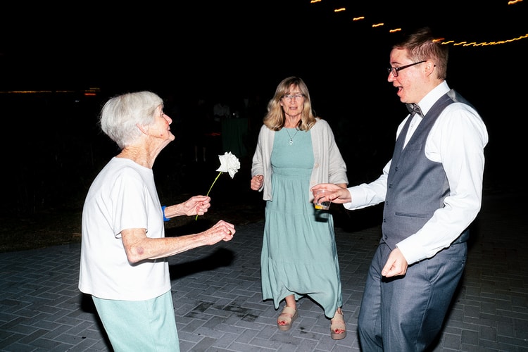 Groom dancing joyfully with elderly and middle-aged family members on the reception dance floor at night, holding a white flower.