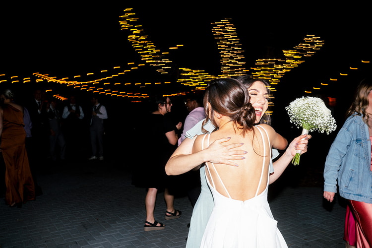 Bride hugging a guest and laughing on the nighttime dance floor, bouquet in hand, streaks of colored light arcing above.