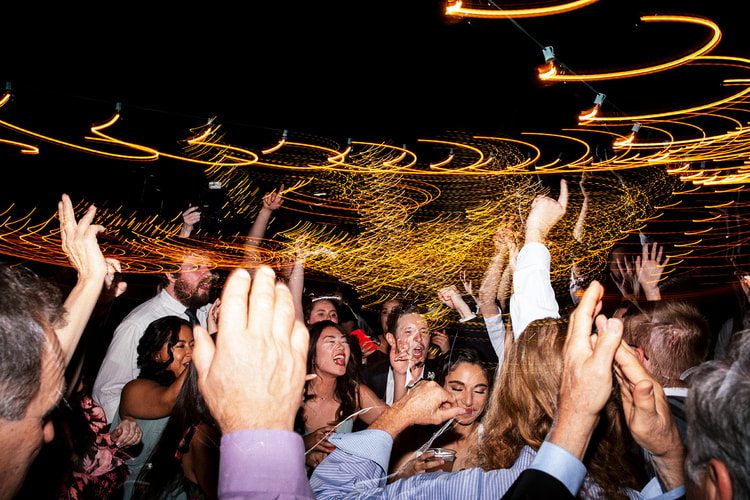 Crowd of guests and couple celebrating on the dance floor, arms raised, surrounded by swirling light trails in the night.