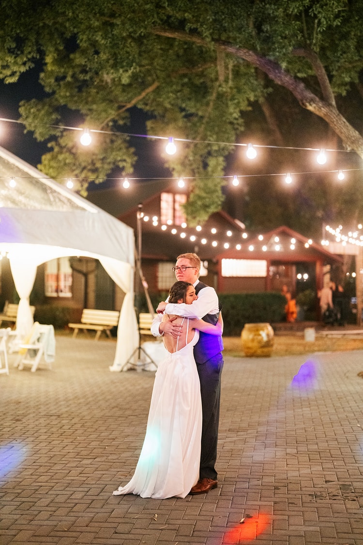 Couple slow dancing outdoors under globe string lights at night, bride's arms wrapped around the groom's neck in a quiet embrace.