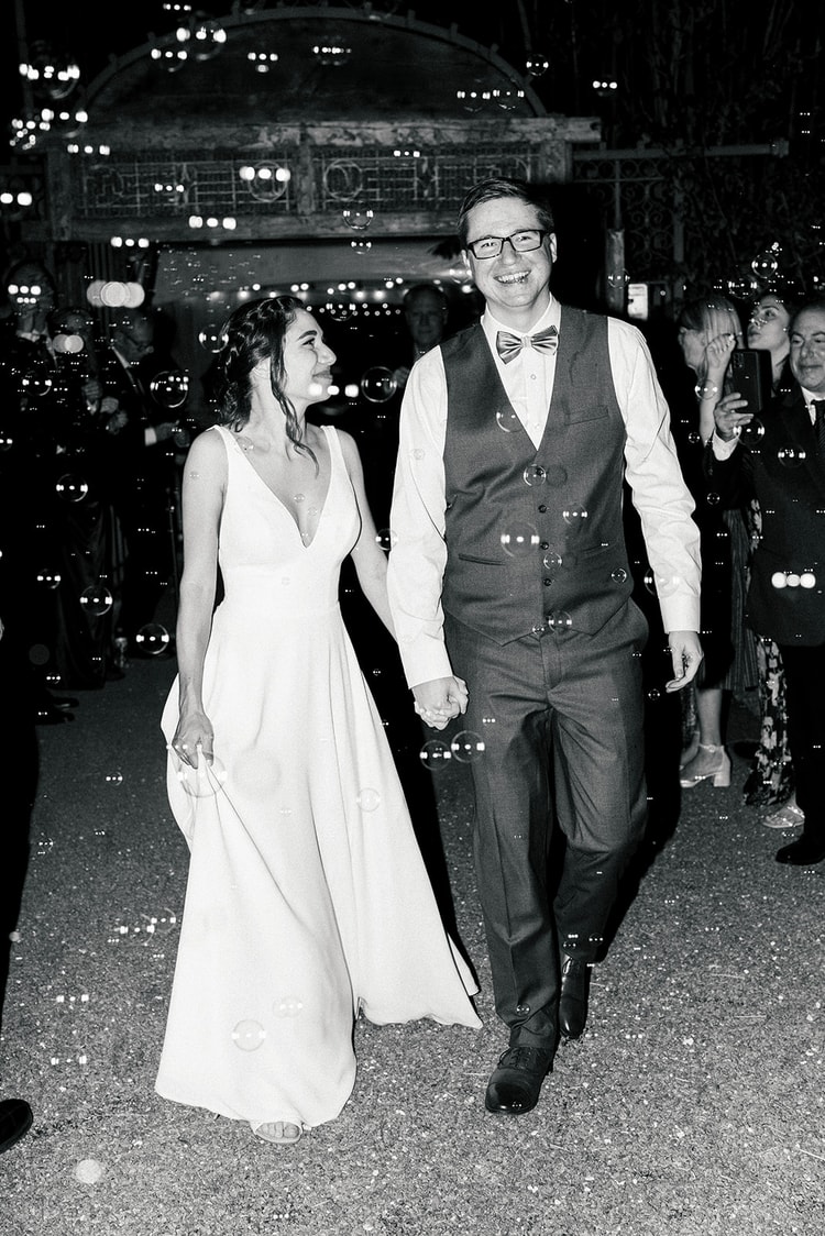 Bride and groom walking hand in hand through a bubble send-off at night, guests cheering on either side, black and white.
