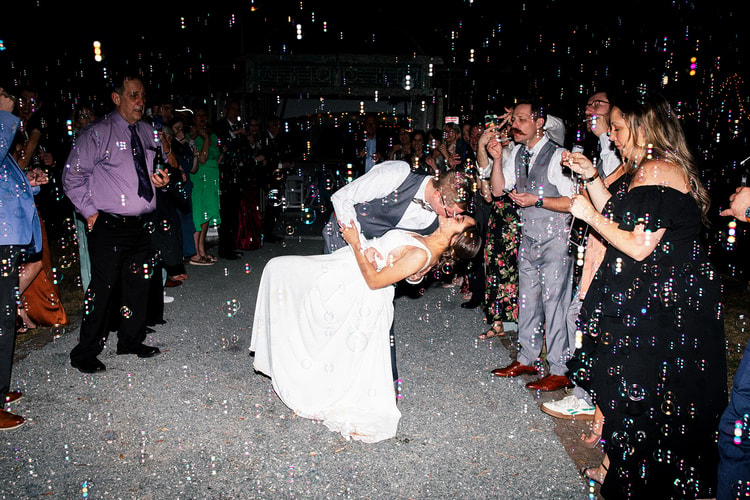 Groom dipping the bride for a dramatic kiss during the bubble send-off, guests cheering and blowing bubbles around them at night.