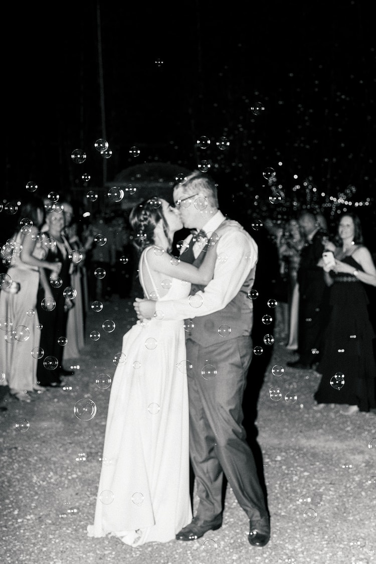 Couple kissing amid a shower of floating bubbles during their send-off, guests watching in the background, black and white.