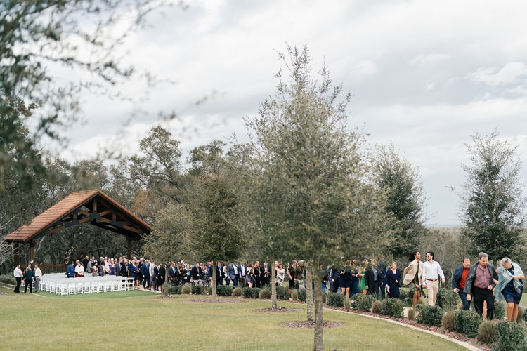 Wedding guests arriving and gathering near an outdoor pavilion ceremony space under a soft, cloudy sky