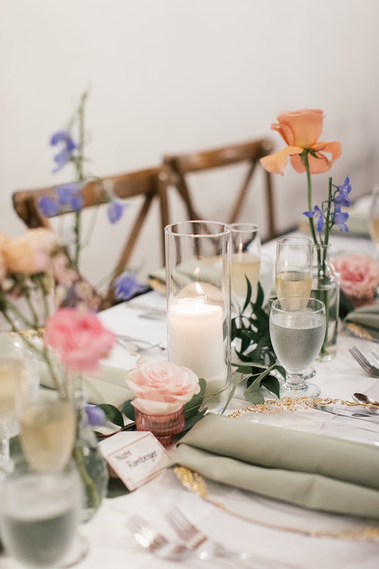 Reception place setting with a sage napkin, gold-rimmed plate, pillar candle, and bud vases of single-stem florals