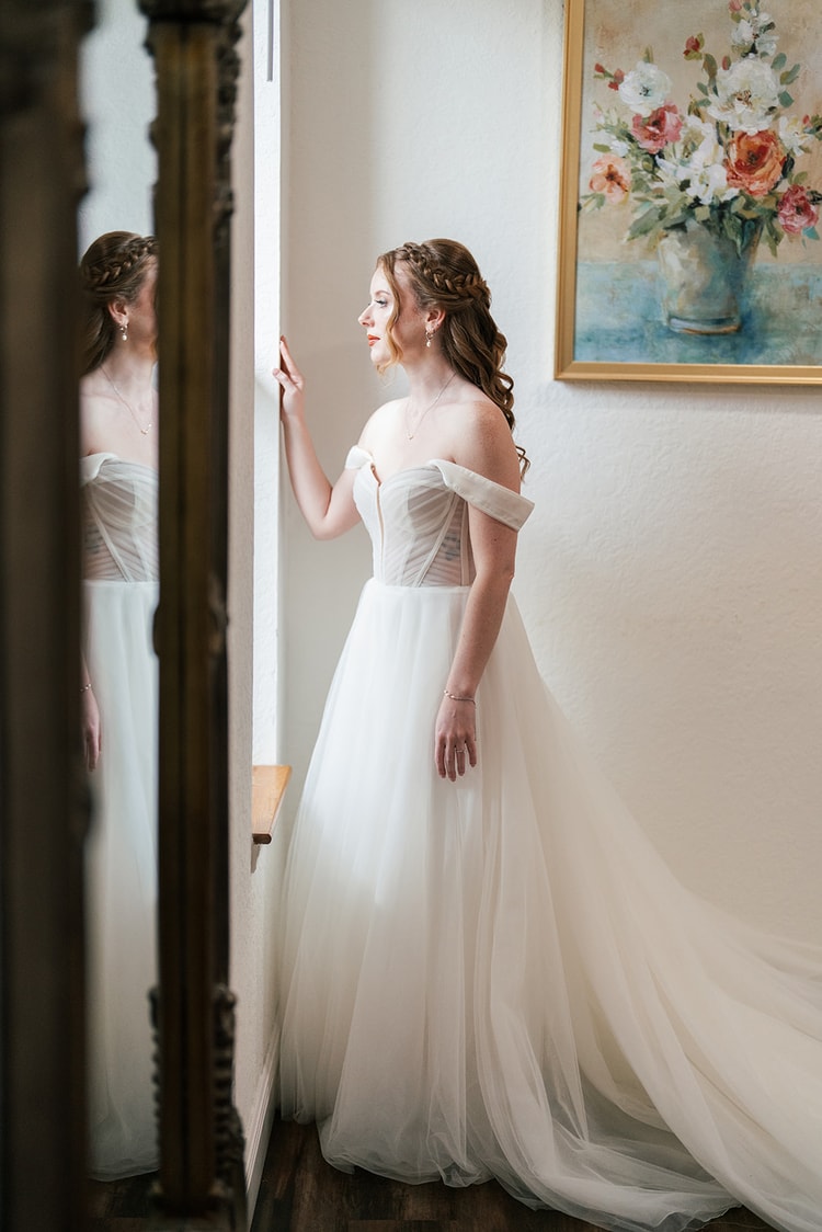 Bride holds her colorful bouquet of pink peonies and orange dahlias close, standing in a bright white hallway.