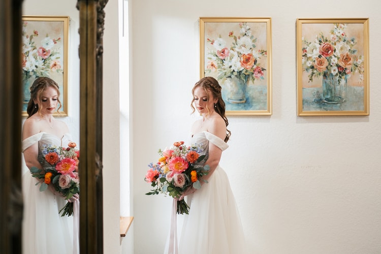 Bride in an off-shoulder gown holds her bouquet while looking down, reflected in a mirror beside floral still-life paintings on a white wall.
