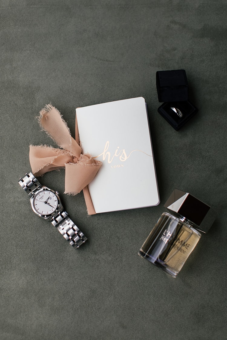 Groom's getting-ready details arranged on sage green fabric: watch, cologne, ring box, and notebook with ribbon