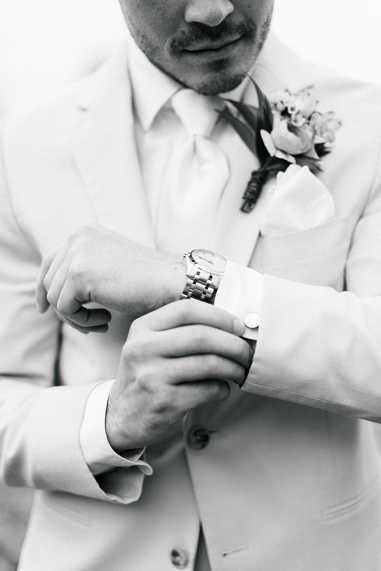 Close-up of the groom's boutonniere featuring a coral ranunculus and blue delphinium pinned to his cream lapel with a white pocket square.