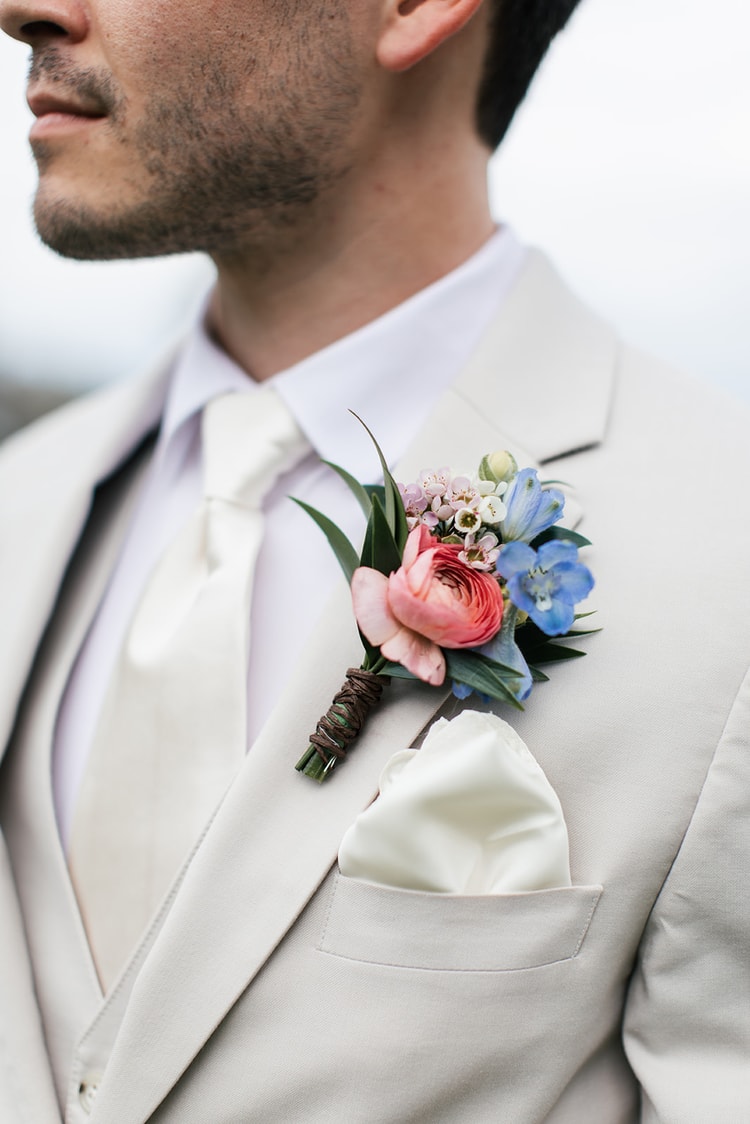 Groomsmen share a laugh together outdoors, one adjusting his cufflink, both dressed in cream suits with floral boutonnieres.