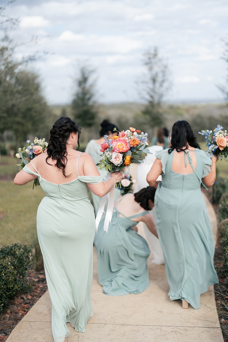 Bridesmaids in sage dresses walk together down a garden path holding bouquets, one bending to adjust the bride's train.