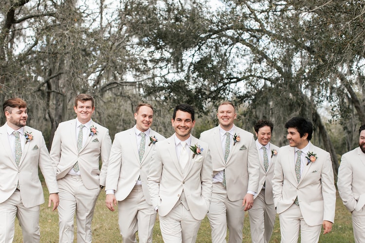 Groomsmen in cream suits and floral ties laugh together under moss-draped oak trees, the groom smiling at the center.