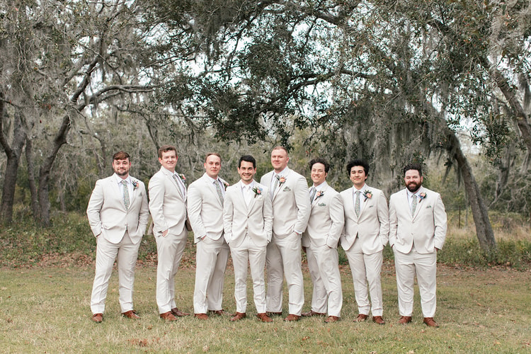 Eight groomsmen in cream suits stand together smiling under sprawling oak trees draped in Spanish moss.