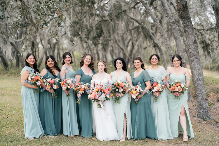 Bride stands with her bridesmaids in sage and green dresses, all holding colorful bouquets under oak trees at the Orlando venue.