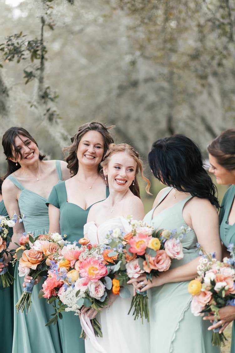 Bride laughs with her bridesmaids gathered close around her, all holding bouquets of coral and peach flowers.
