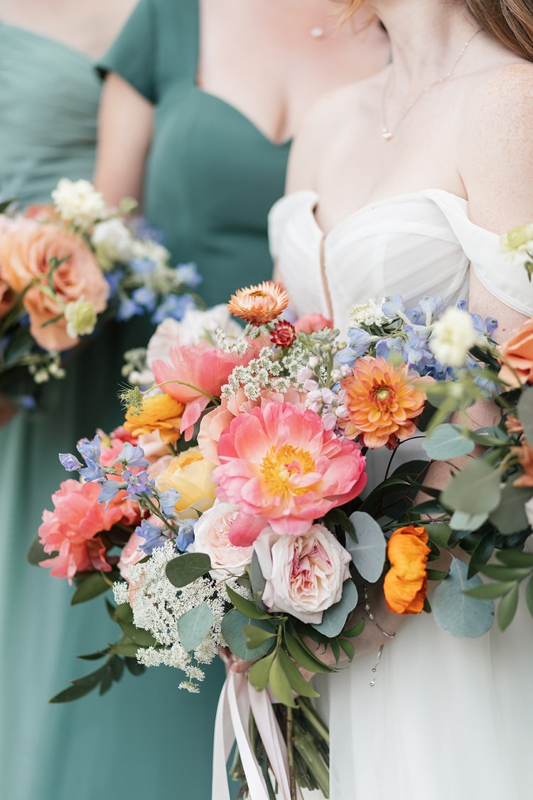 Bride holds a bright bouquet of coral peonies, orange dahlias and blue delphinium against her off-shoulder gown, bridesmaids in sage green beside her.
