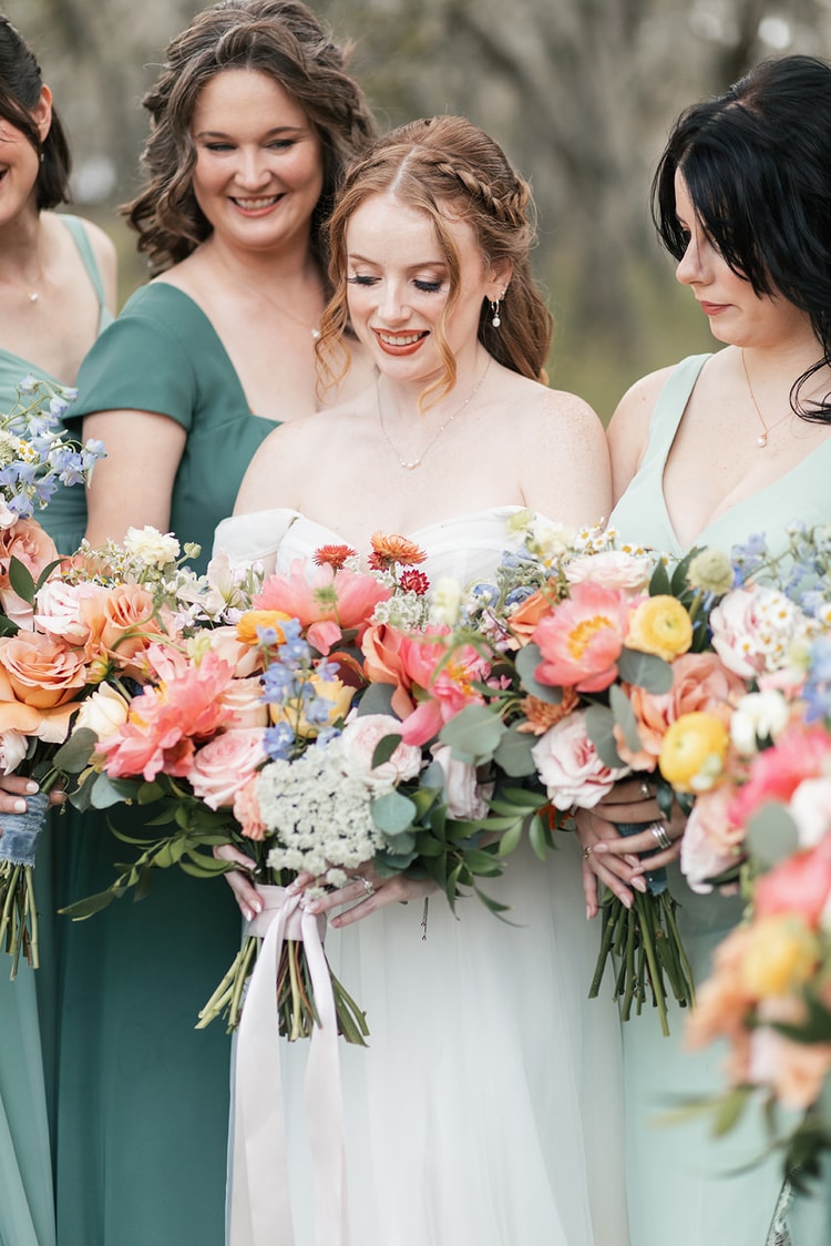 Bride smiles among her bridesmaids as they hold their bouquets together in a close huddle outdoors.