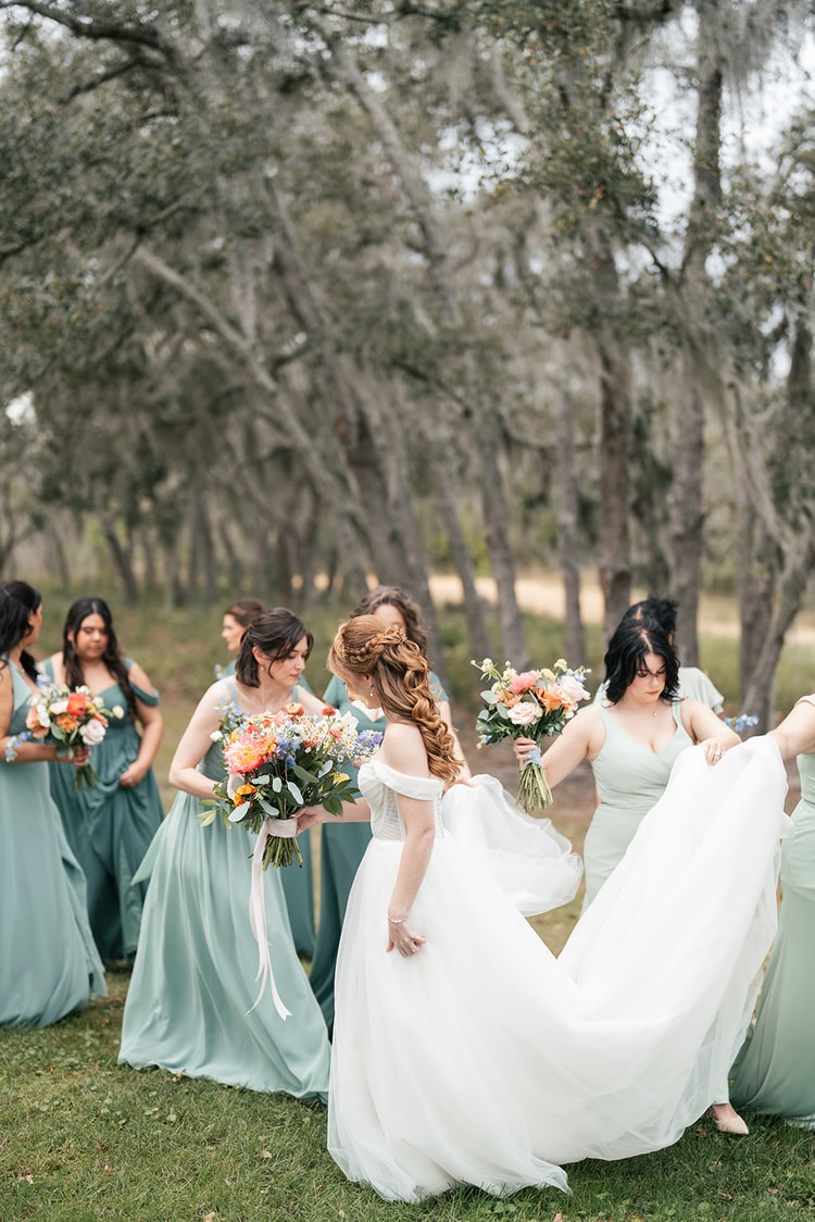 Bridesmaids in sage dresses walk along a path with their bouquets as one helps arrange the bride's flowing gown.