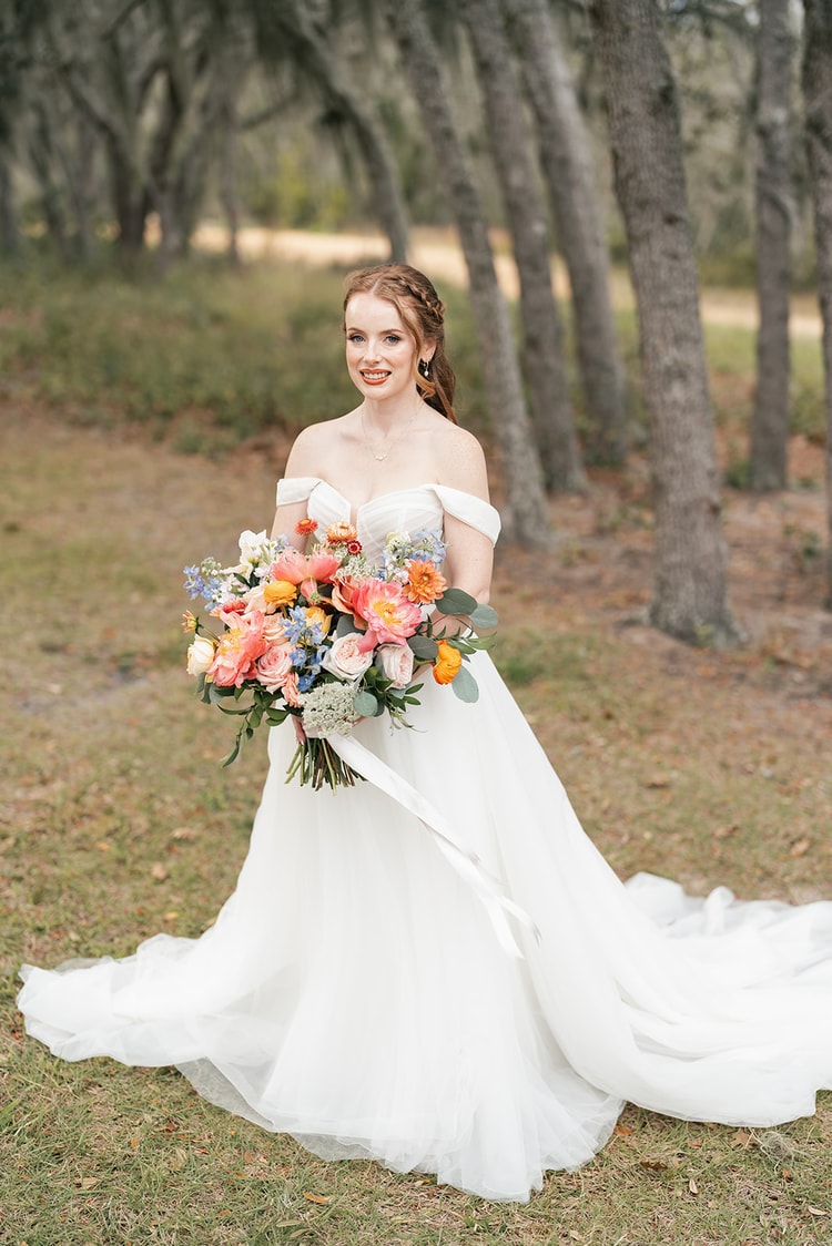 Bride smiles while holding her bouquet of pink peonies and orange dahlias, standing among oak trees with her train spread on the grass.