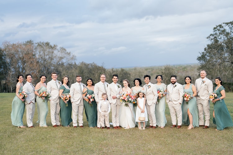 Full wedding party in sage green and tan attire poses together on the lawn with rolling countryside in the background.