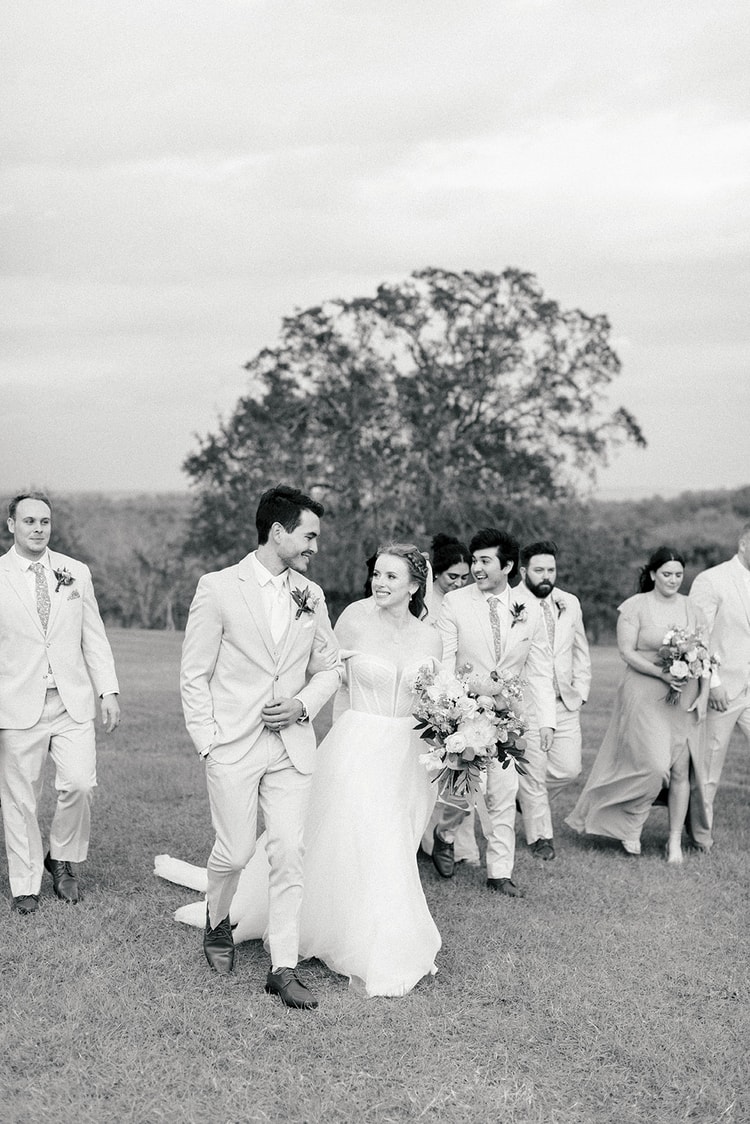 Black and white photo of the newlyweds walking arm-in-arm across an open field with their wedding party following behind.