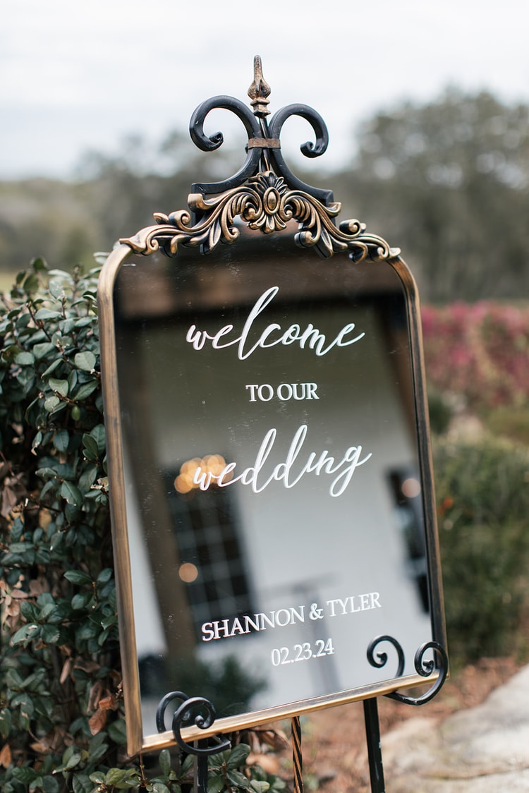 Mirrored welcome sign reading Welcome to Our Wedding, Shannon and Tyler, 02.23.24, framed by greenery at the venue entrance.